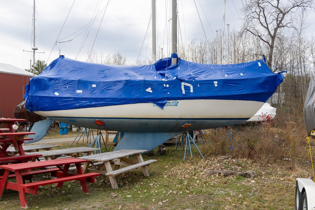 A sailboat covered with a blue shrinkwrap and white hull, supported by stands outdoors in a yard with picnic tables, bare trees, and an overcast sky.