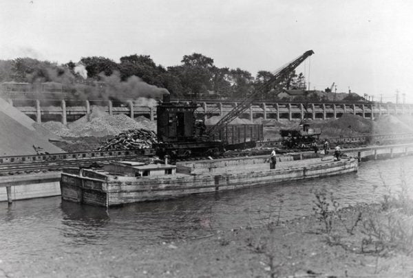 Historical black and white photograph of a steam-powered crane on a barge near a dock with trees in the background. Now the location of Bridgeview Harbour Marina.